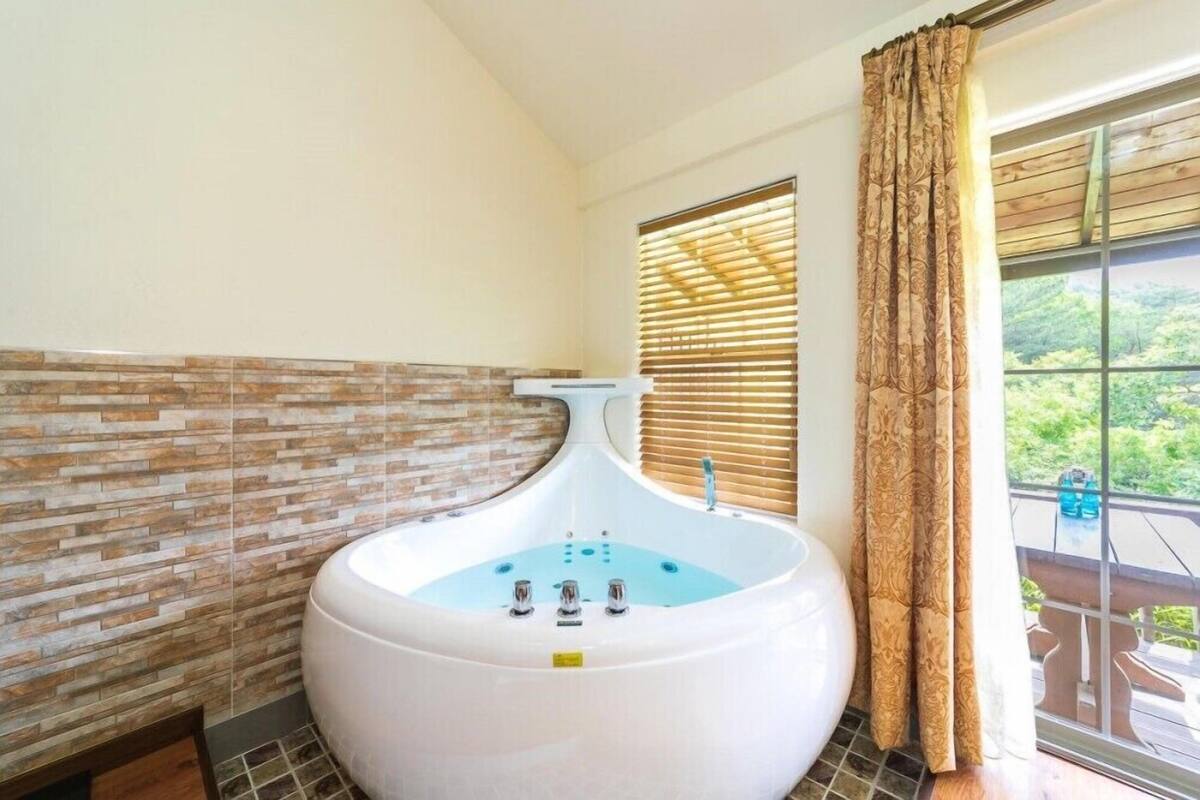 A spa room with a large bathtub, a window with blinds, and a view of greenery outside.
