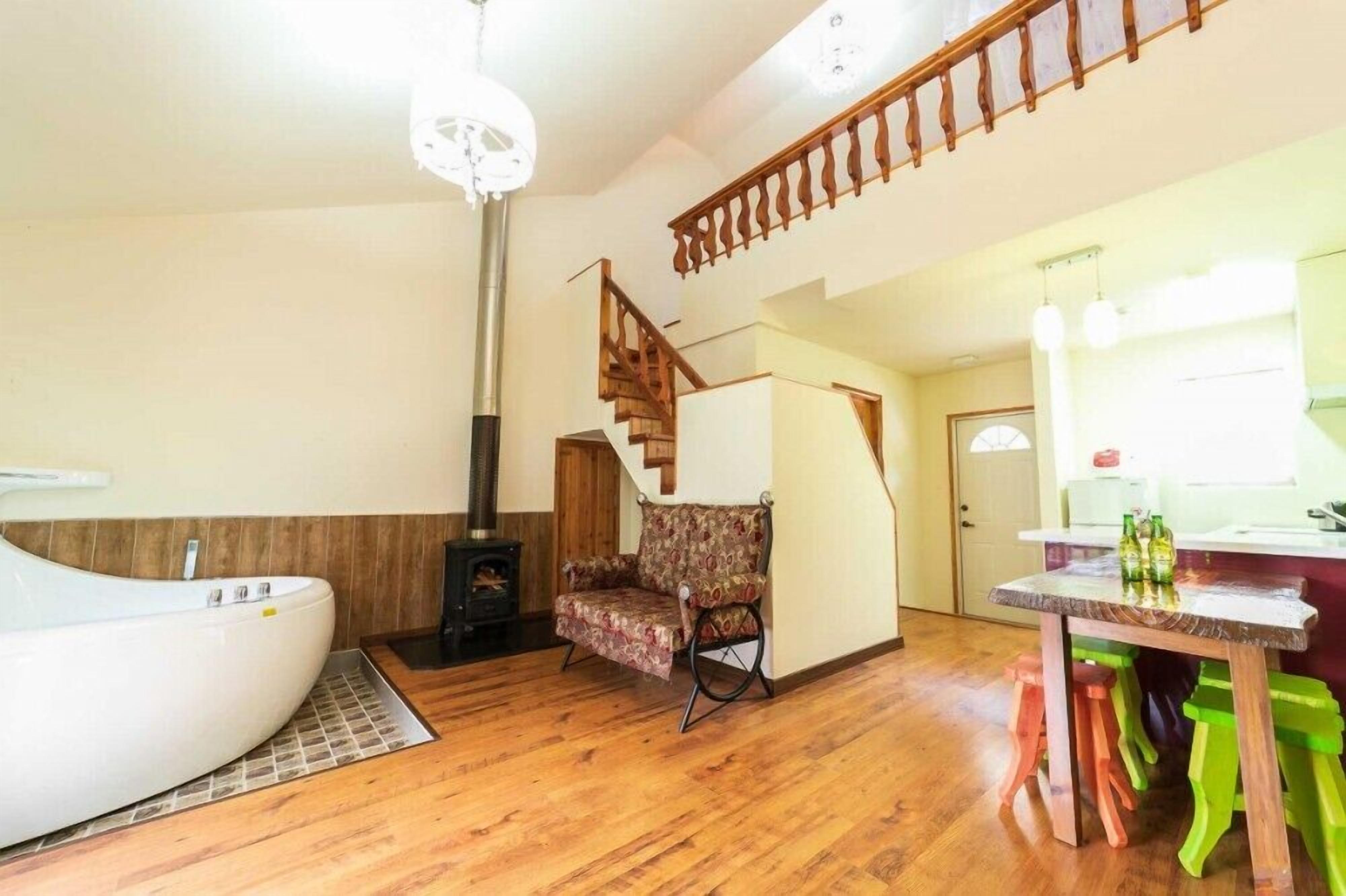Amplia sala de estar con piso de madera, una bañera blanca, un sillón estampado, una escalera de madera y una zona de cocina con una mesa y taburetes.