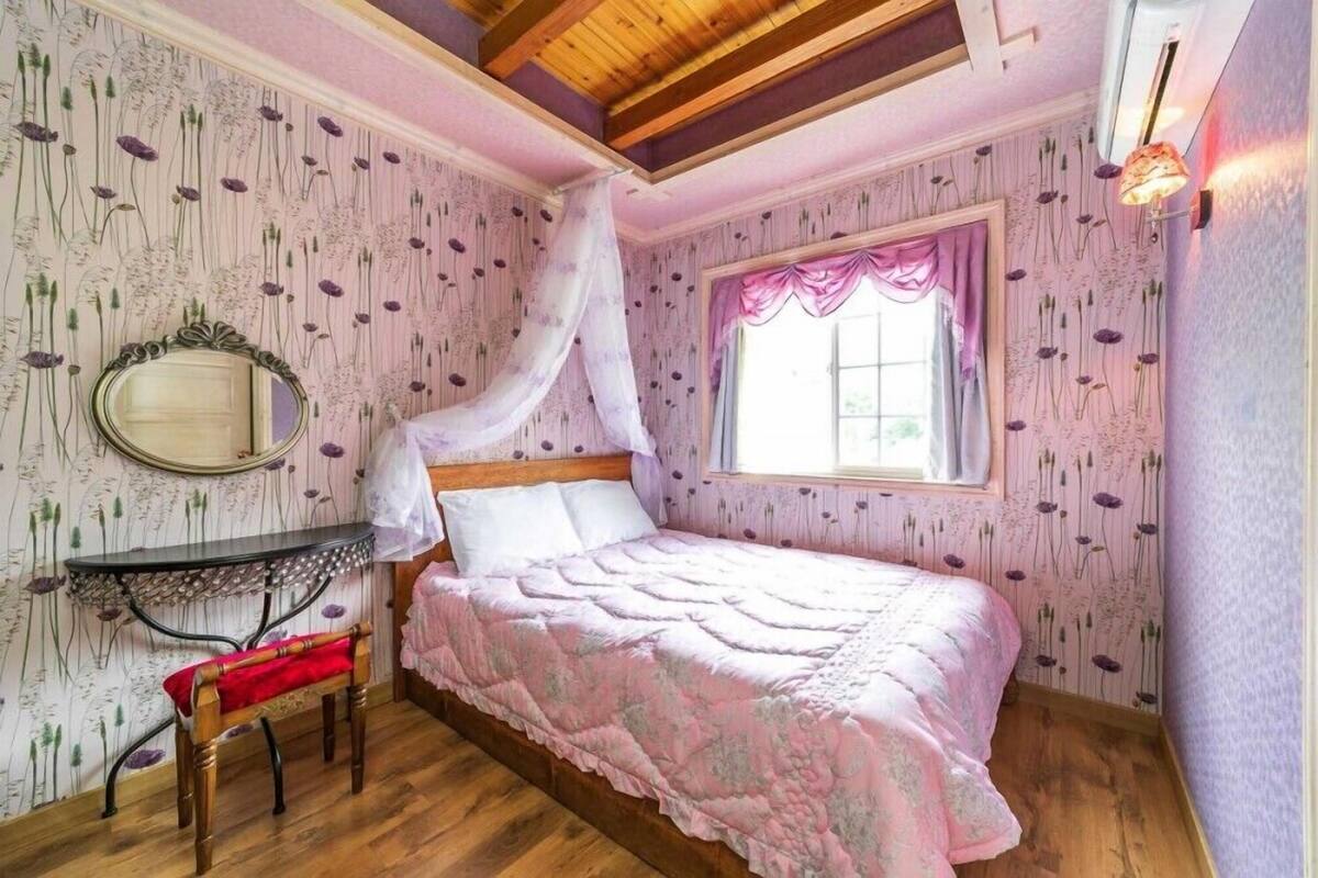A bedroom with a bed, a bedside table, a mirror, a window with a curtain, and floral wallpaper.