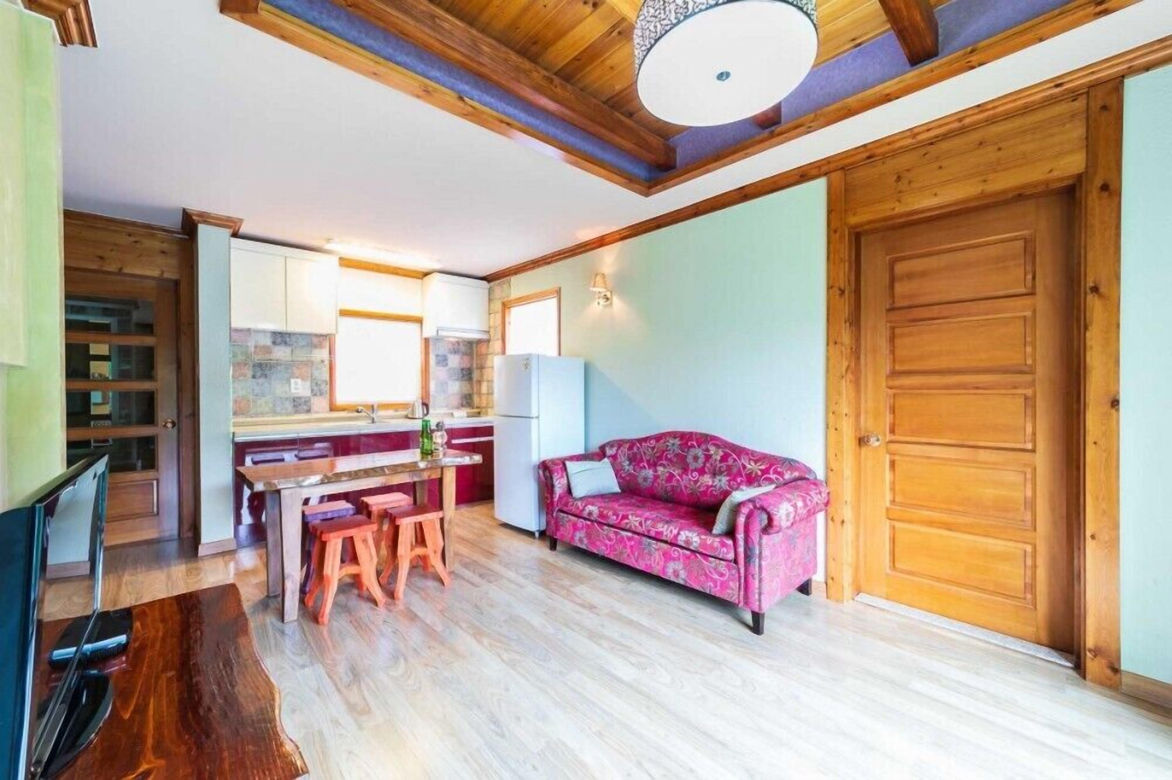 A cozy living room with a wooden ceiling, a pink patterned sofa, and a dining area with a wooden table and stools.