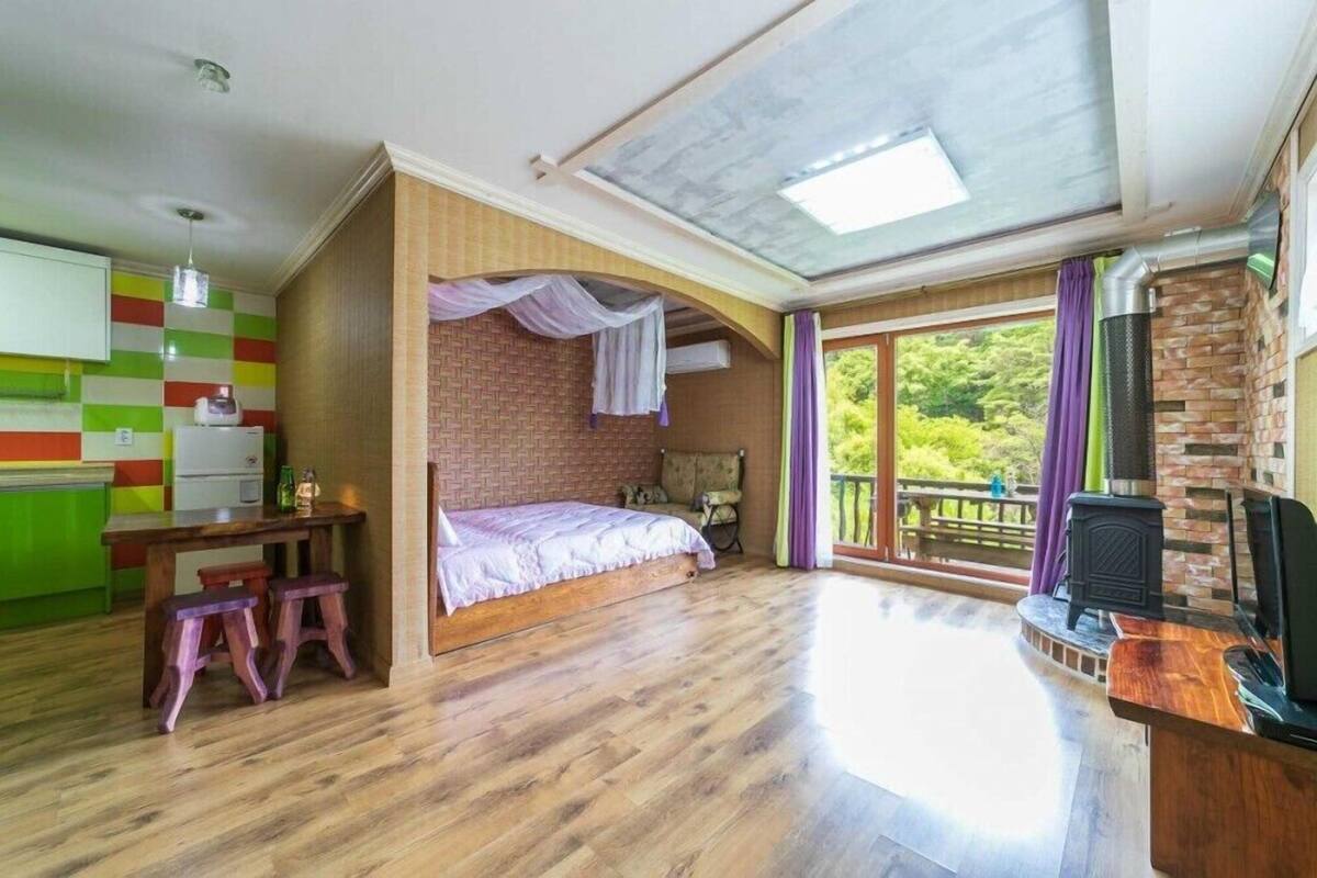 A bedroom with a bed, a wooden desk, a chair, and a window with a view of greenery.
