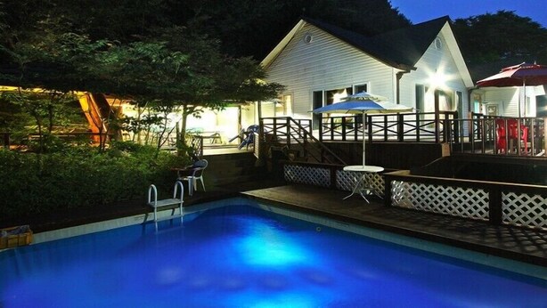 A swimming pool with a wooden deck, a covered patio area with umbrellas, and a white building with a porch. - Window to the sky Pension (Gapyeong)