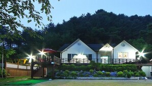 A row of three white cabins with a wooden deck and a small bridge connecting them, surrounded by greenery and a forested backdrop. - Window to the sky Pension (Gapyeong)