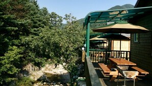 Un área de comedor al aire libre con mesas y bancos de madera, una zona de estar cubierta con sombrillas, y una vista de un río y montañas boscosas al fondo.