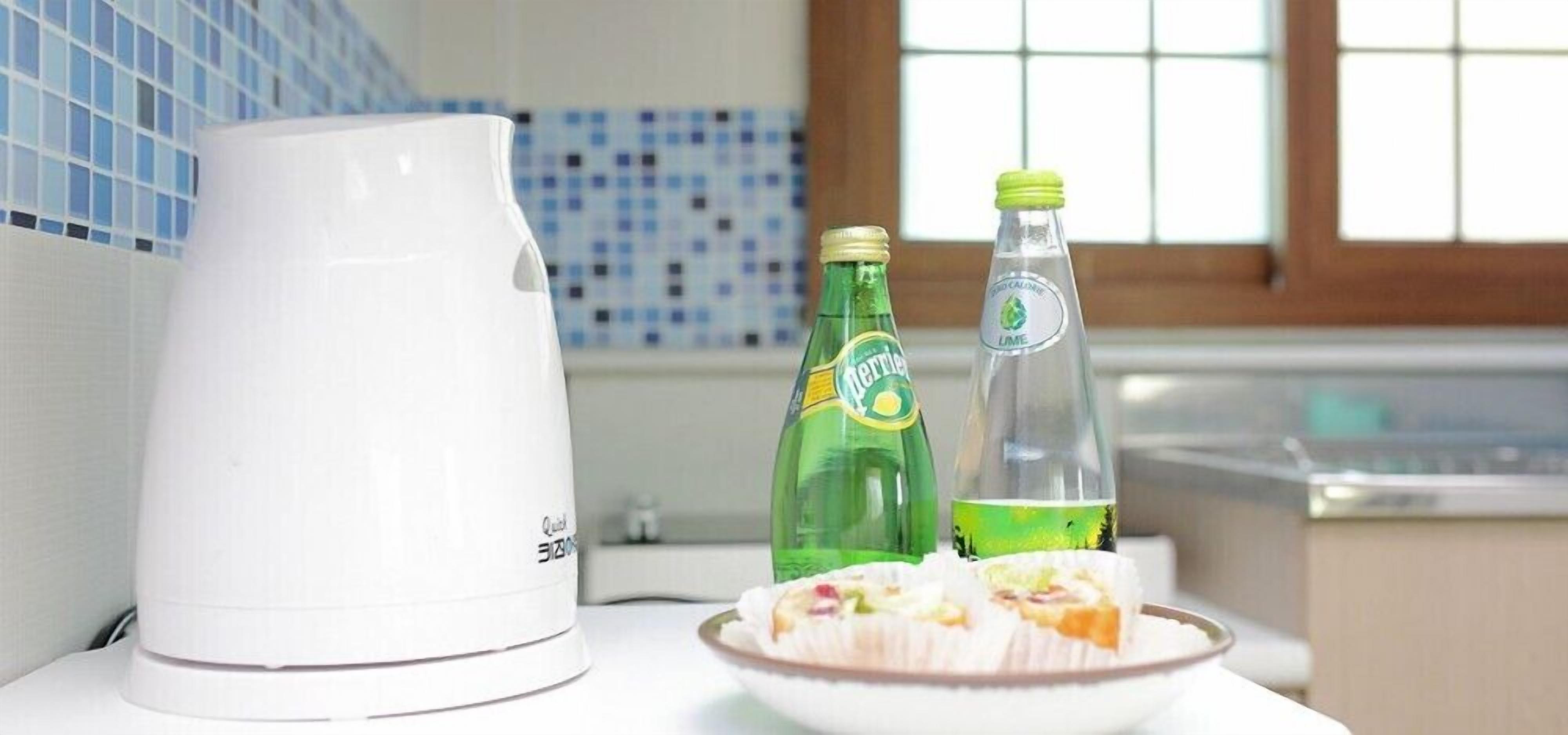 A white kettle, two bottles of lime soda, and a plate with food on a table in a kitchen with blue and white tiles.