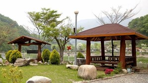 A gazebo with a wooden roof and bench, surrounded by rocks and shrubs, set in a landscaped garden with trees and a mountain in the background. - Bandi Hwangto Pension (Miryang)