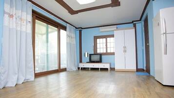Una habitación con suelo de madera, un refrigerador blanco, un armario blanco, un televisor sobre una mesita y una ventana con cortinas.