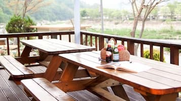 Una mesa de picnic de madera con bancos, una cesta de manzanas y botellas en un porche con vistas a un jardín y árboles.