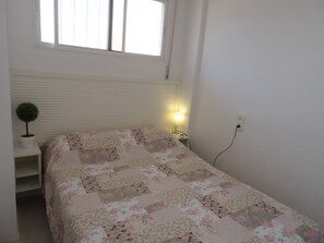 Une chambre à coucher comprenant un lit, une table de chevet, une lampe, une prise de courant et une fenêtre avec des stores.