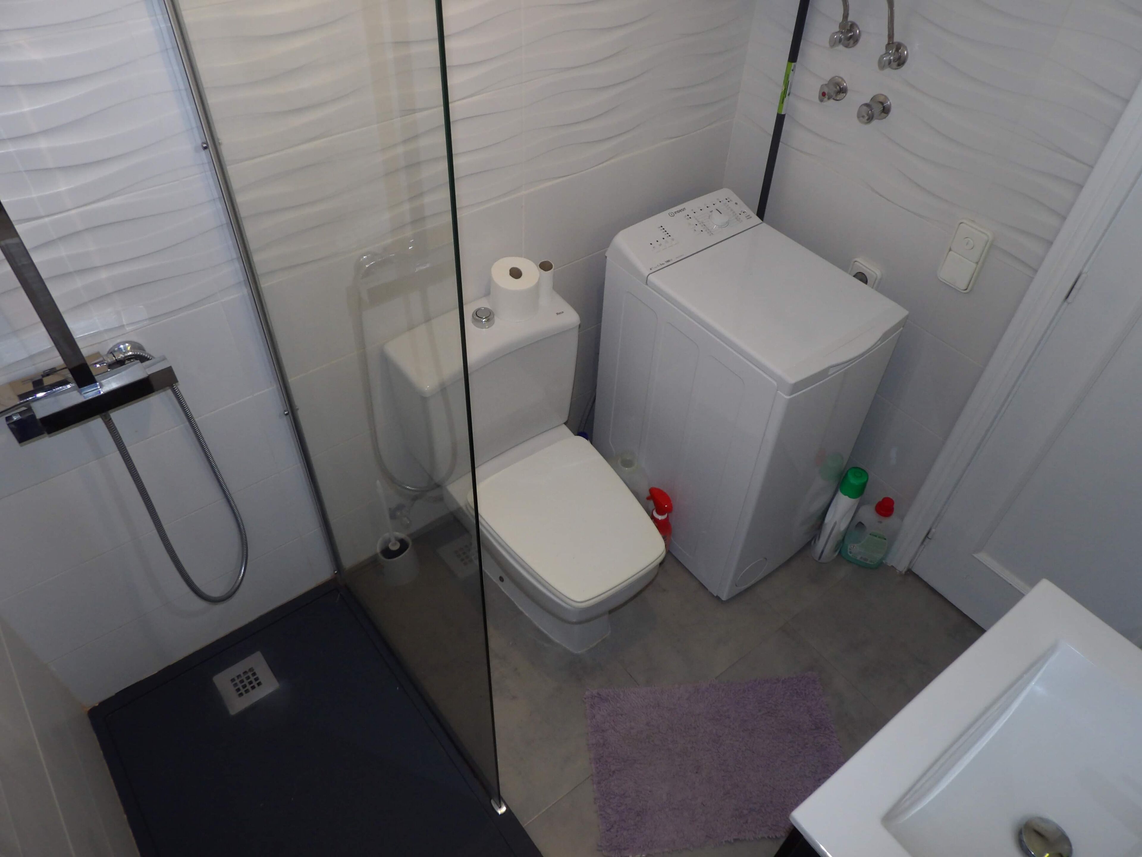 A compact bathroom with a glass shower enclosure, a white toilet, and a compact washing machine.