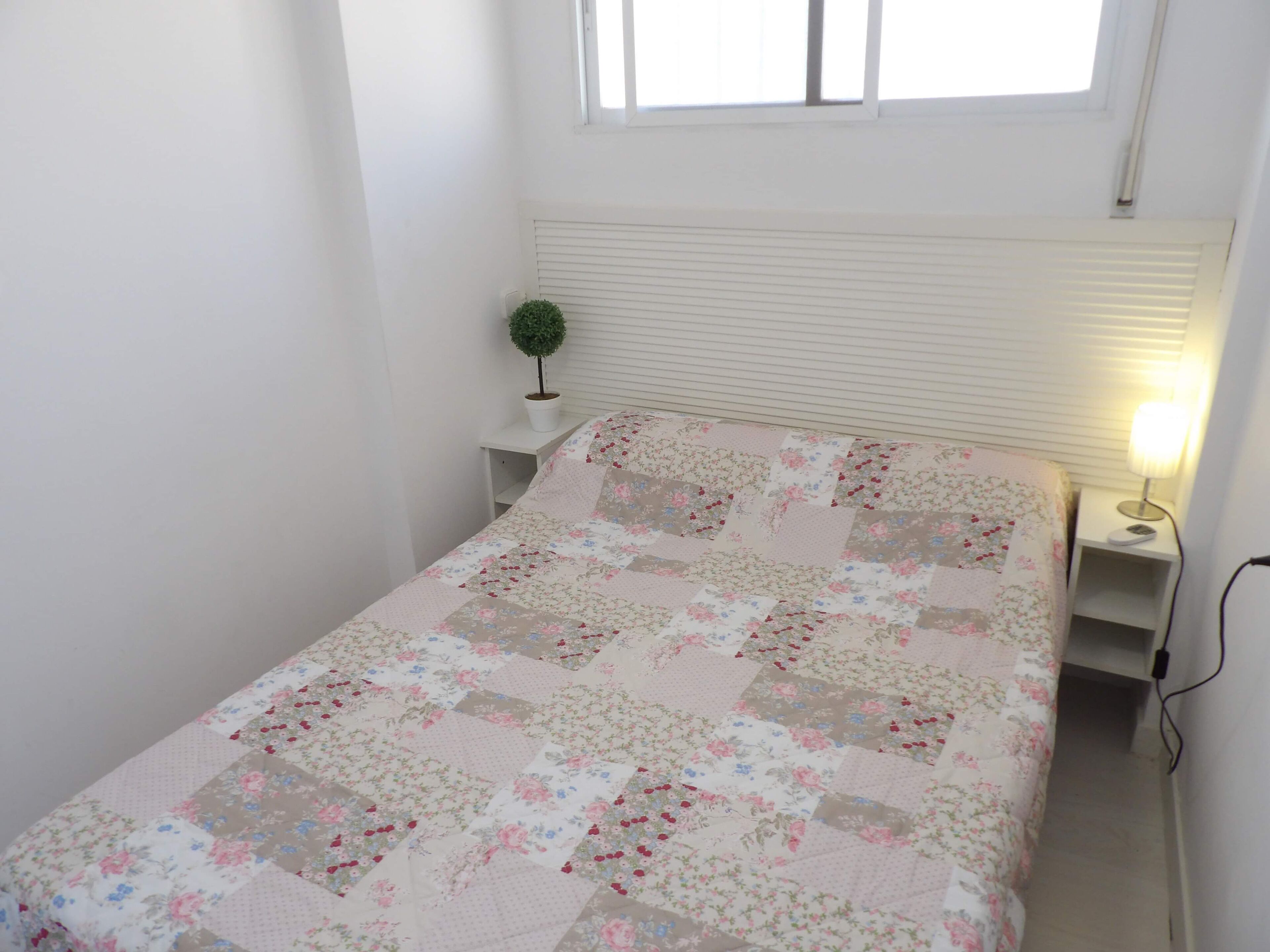 A small, neatly arranged bedroom with a floral quilt, a bedside lamp, and a small potted plant.