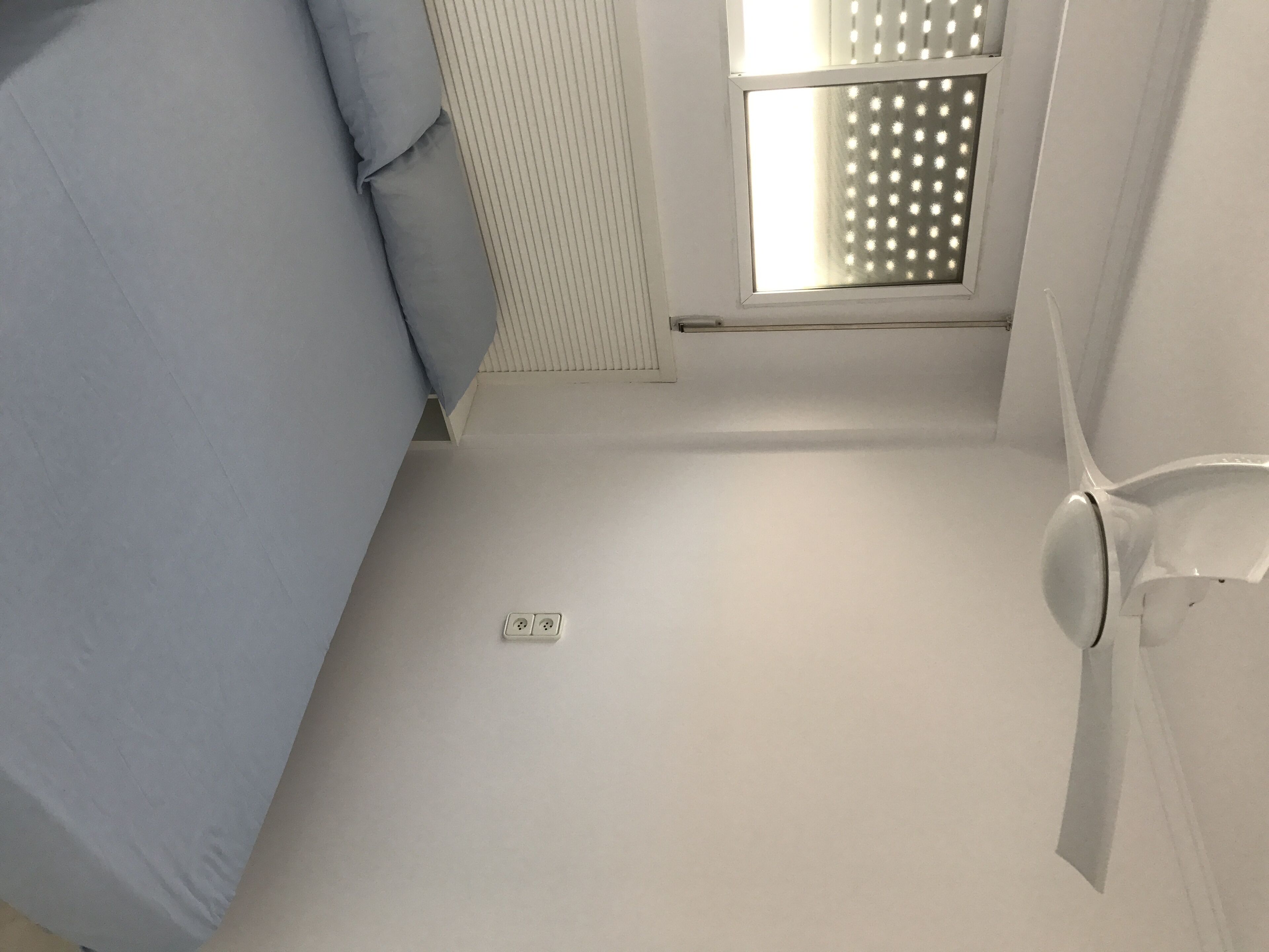 A small, sparsely furnished room with a single bed, a window with blinds, and a visible electrical outlet.