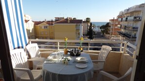 Un balcó amb una taula rodona preparada per a dues persones, dues cadires de plàstic blanc i vista al mar i els edificis.