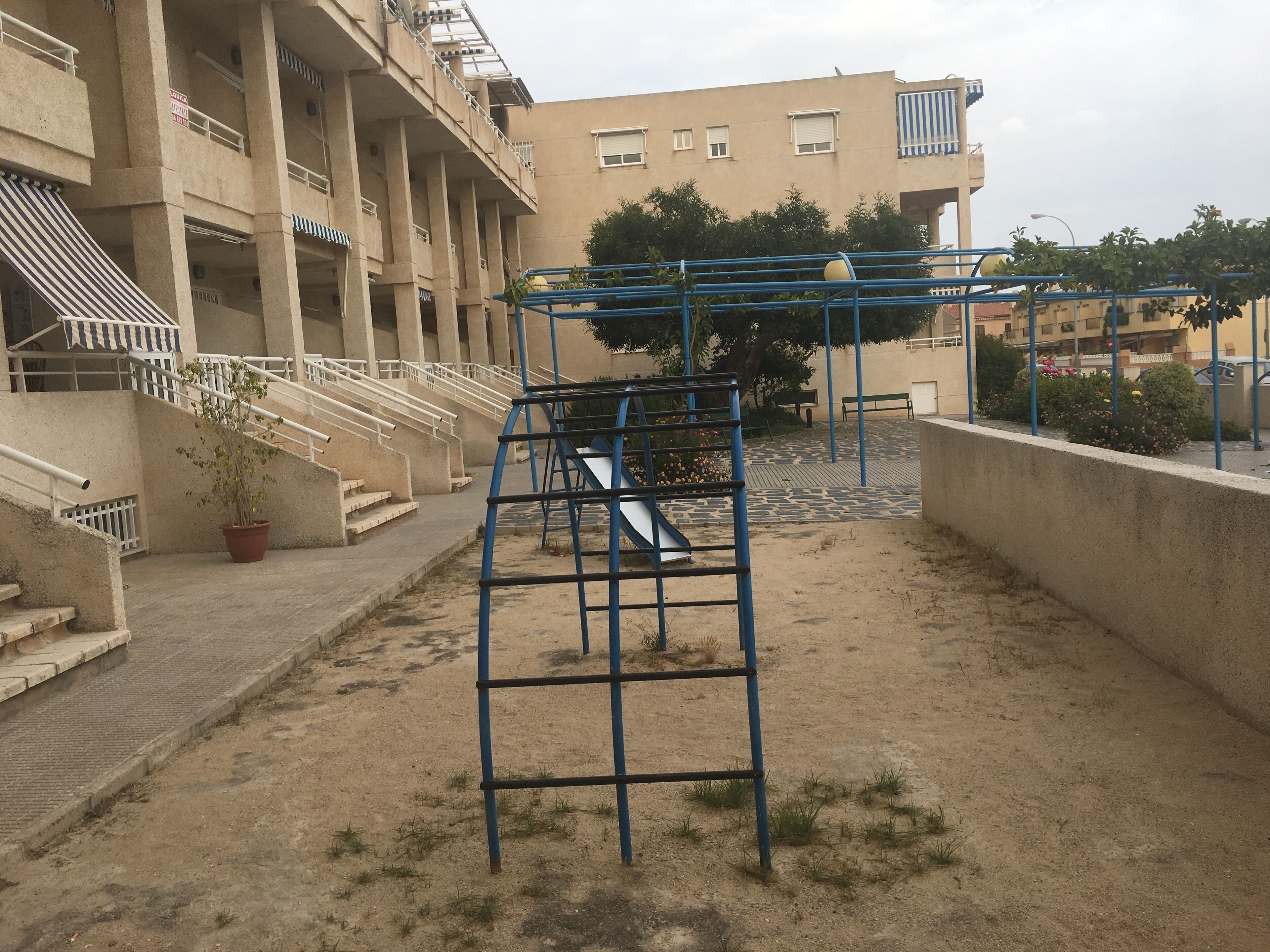 Un parque infantil con juegos azules, una escalera y un edificio de varios pisos al fondo.