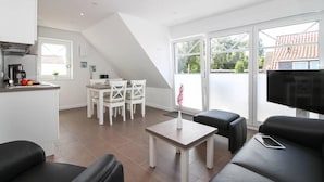 Una sala de estar moderna con un sofá, una mesa de centro y un televisor. Hay un comedor con una mesa y sillas. La habitación tiene ventanas amplias y vistas al exterior.