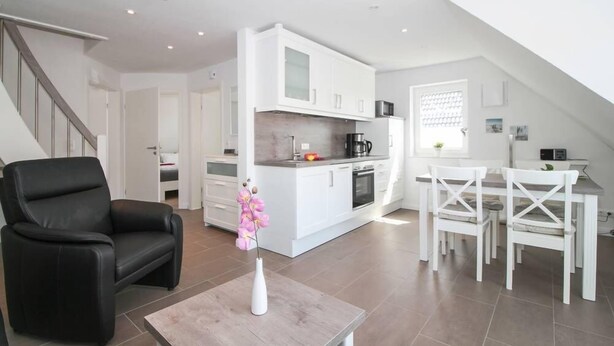 Una cocina moderna con armarios blancos, un comedor con sillas blancas y un sillón de cuero negro.