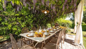 Un área de comedor al aire libre con una mesa dispuesta para una comida, rodeada de abundante vegetación y flores moradas.