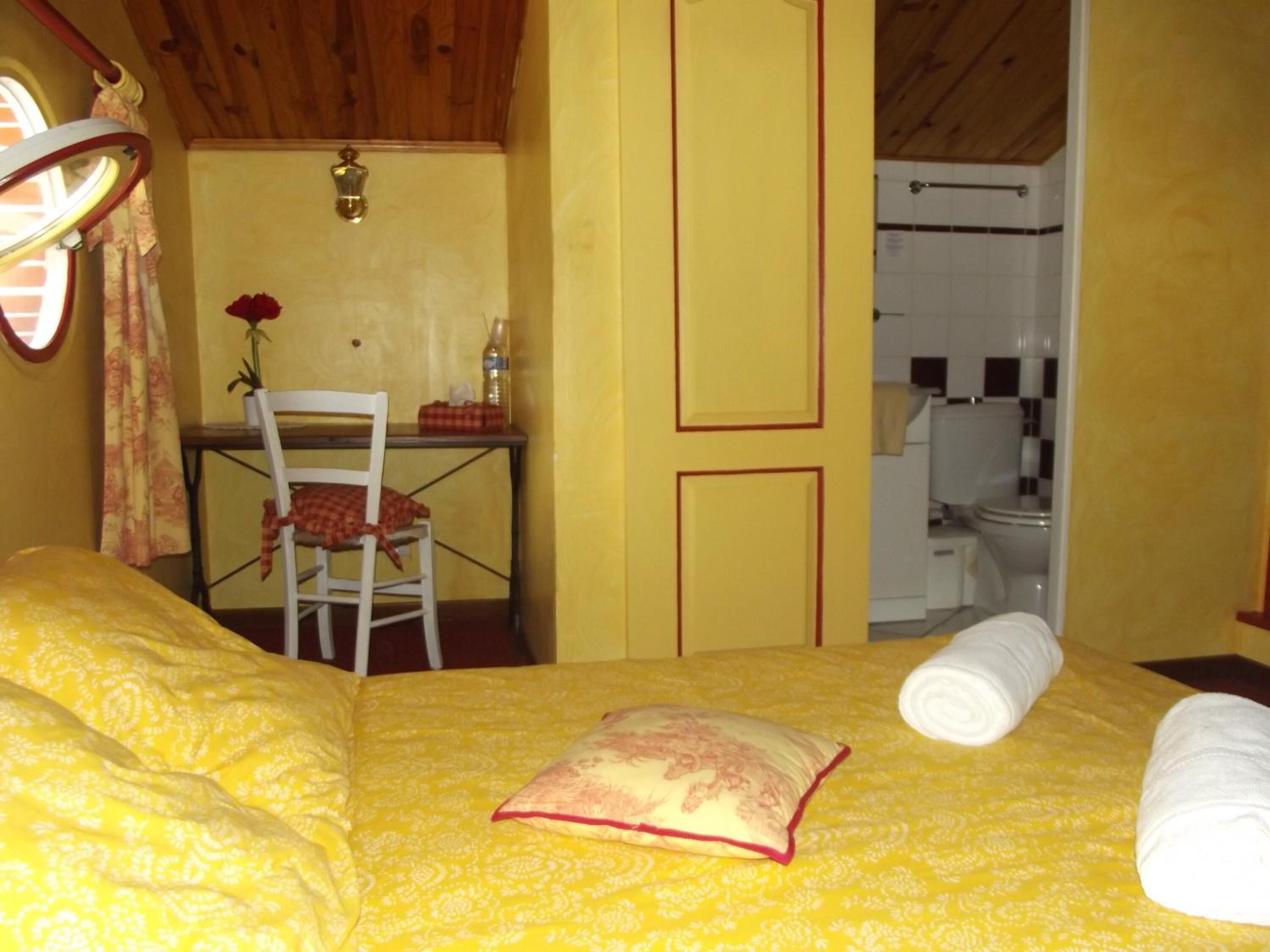 Une pièce comprenant un lit, une chaise, une petite table avec une bouteille et une fleur, et donnant sur une salle de bain.