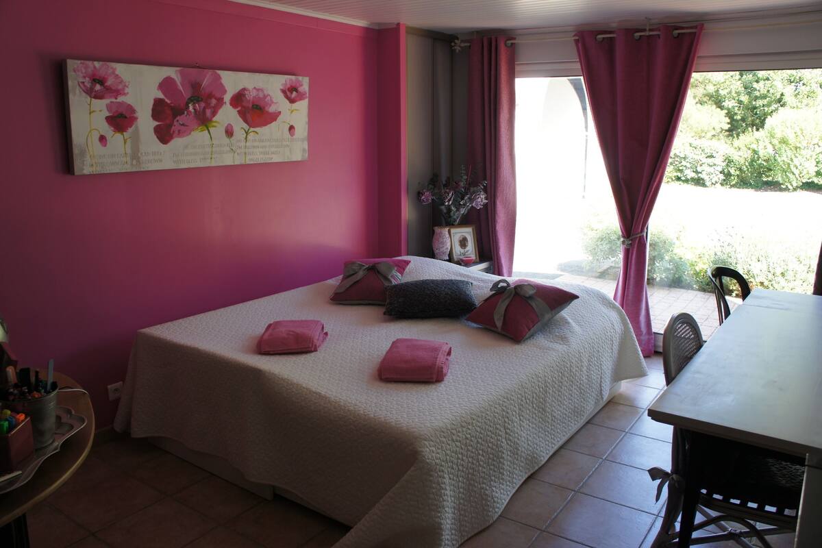 Ein Zimmer mit einem Bett, einem Tisch und einem Stuhl. Ein Fenster mit Vorhängen, eine Vase mit Blumen und ein Gemälde an der Wand.