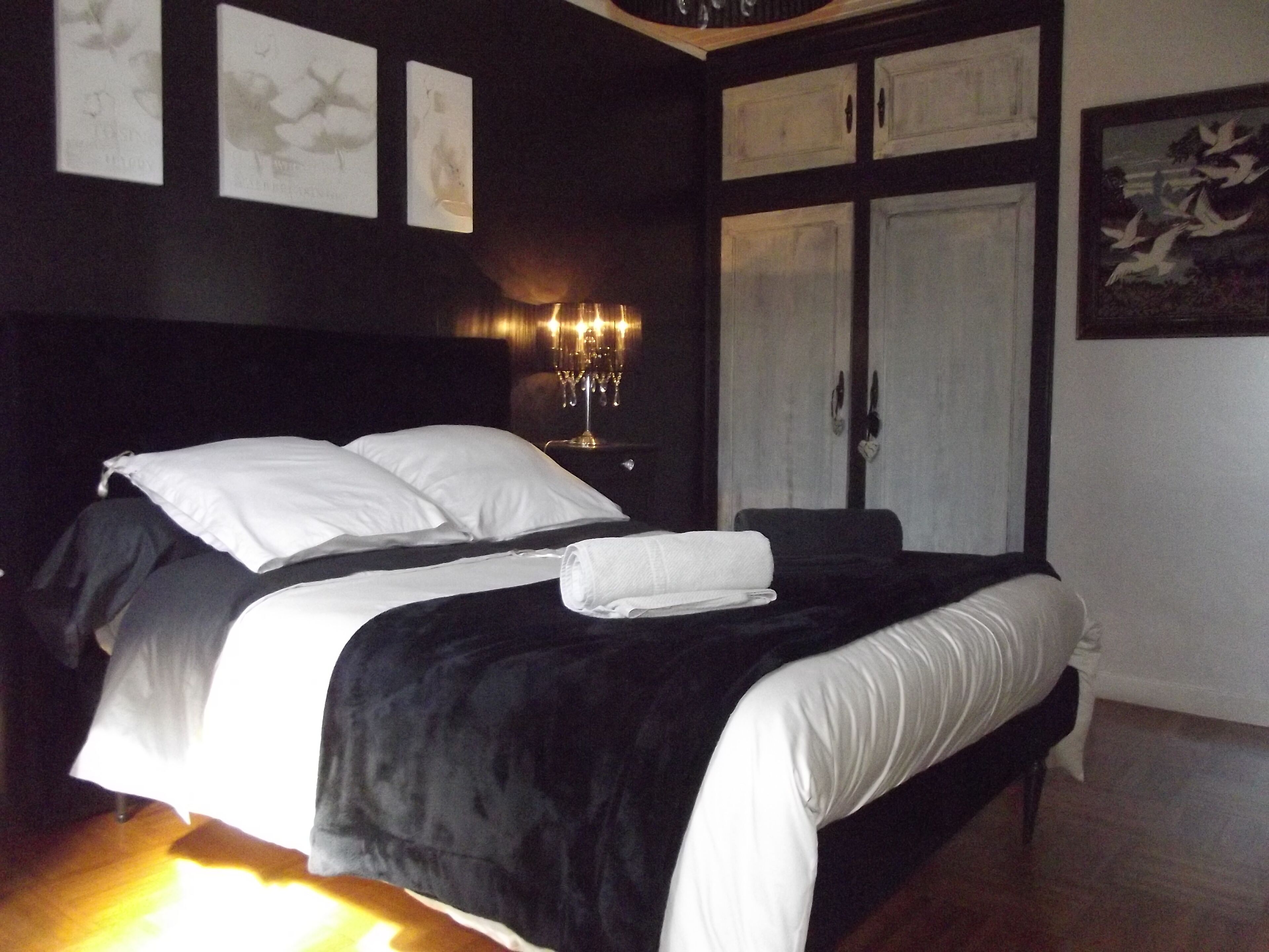 Un lit bien fait, avec du linge de lit blanc, une tête de lit noire et une table de chevet surmontée d’un chandelier.