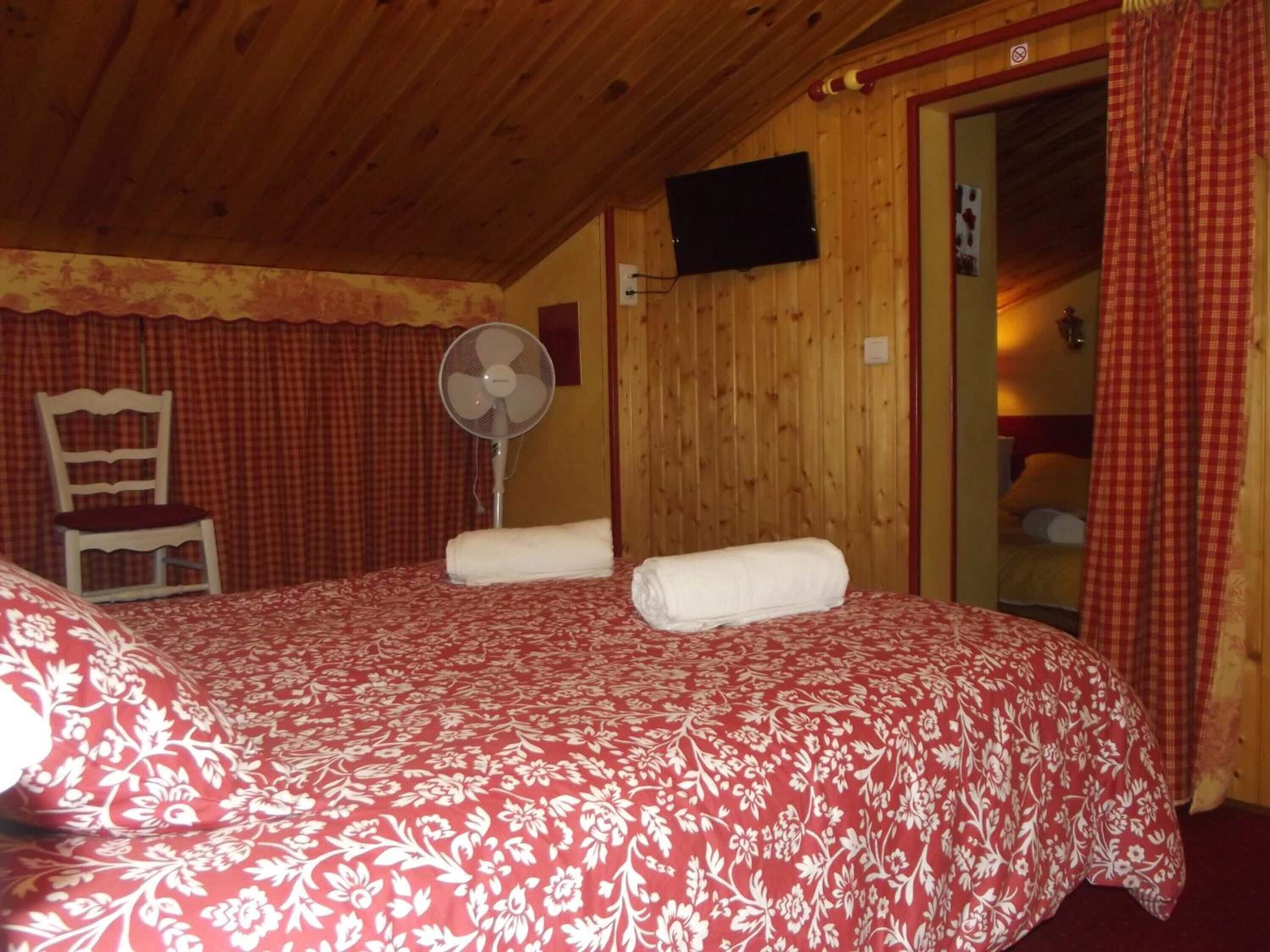Un cuarto acogedor con techo de madera, una cama con colcha floral roja, una silla blanca, un ventilador de pie y un televisor montado en la pared.