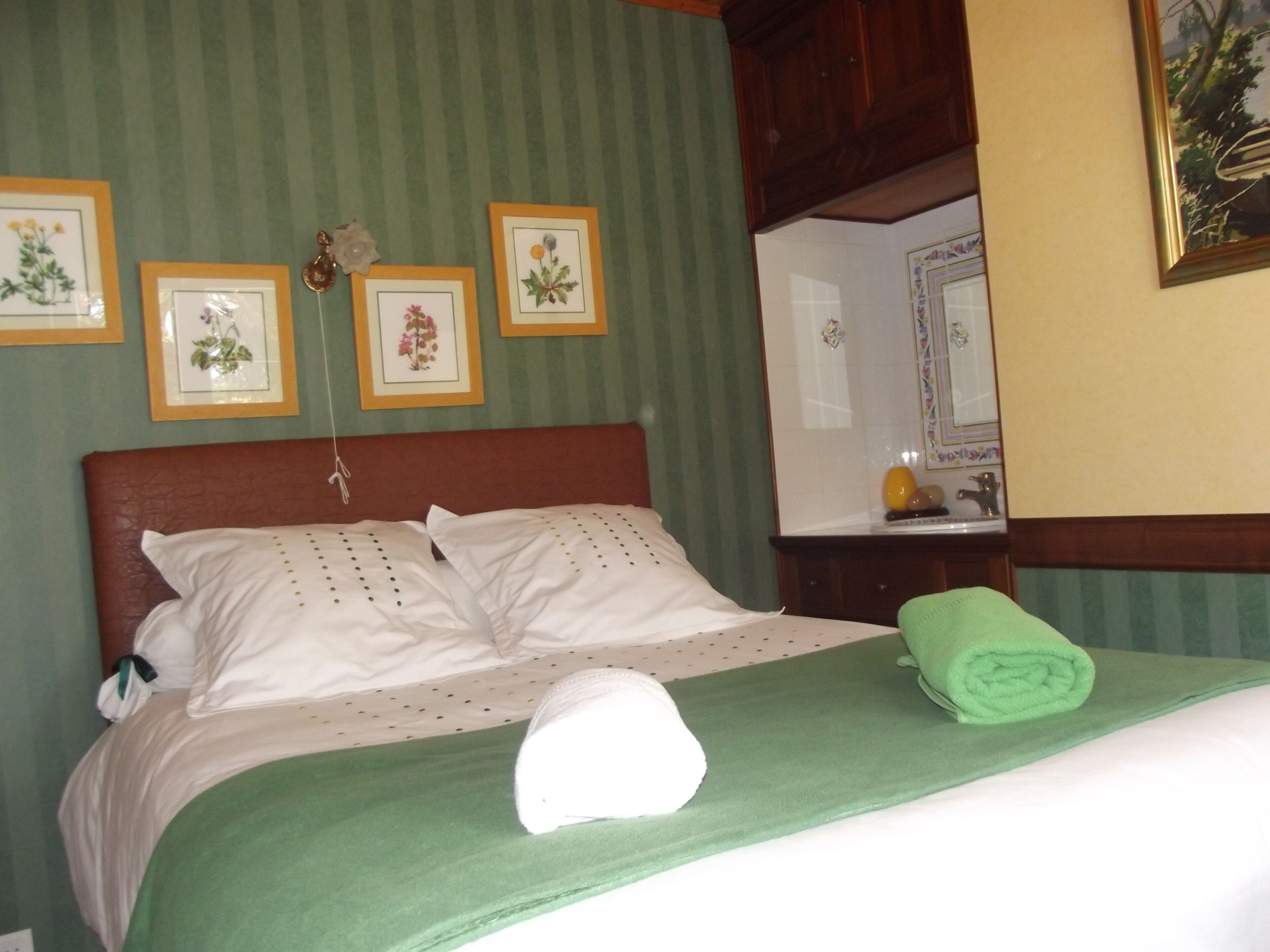 A neatly made bed with a green bedspread, white pillows, and a rolled green towel. There are four framed botanical prints on the wall above the bed.