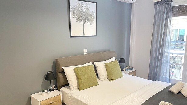 A neatly arranged bedroom with a bed, bedside tables, a nightstand, a wall-mounted air conditioner, and a window with curtains. - Check Point - Veikou (Athens)