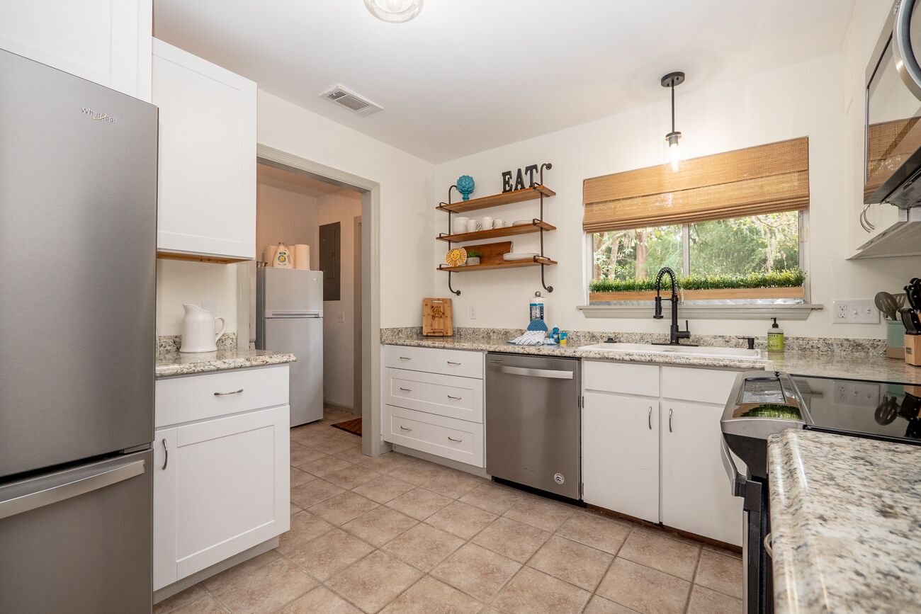 A modern kitchen with white cabinetry, a stainless steel refrigerator, a dishwasher, and a granite countertop. There are wooden shelves above the sink and a window with a view of greenery.