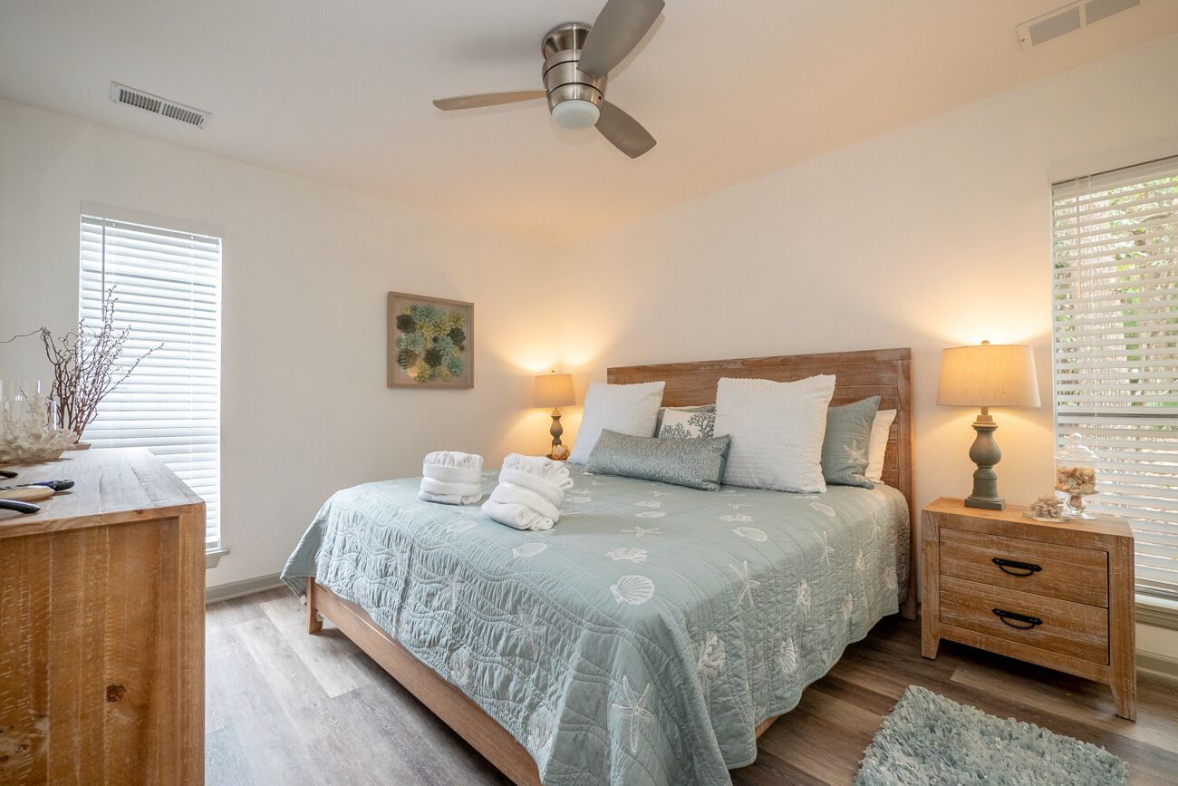 Une chambre à coucher comprenant un lit, des tables de chevet, un ventilateur de plafond et une fenêtre avec des stores.