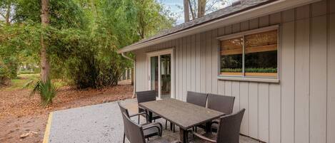 An outdoor patio with a table and chairs, surrounded by trees and a building with a window.