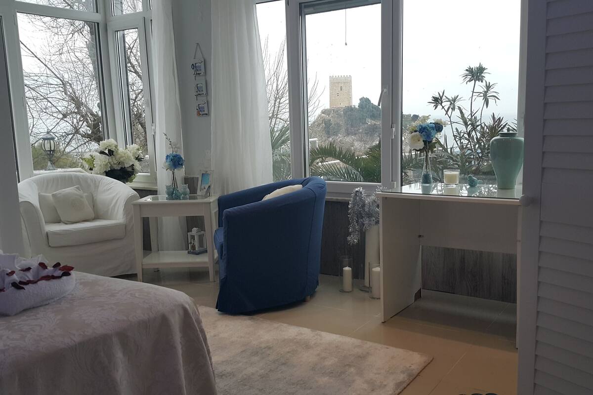 A room with a bed, a white armchair, a blue armchair, a small table with a vase of flowers, and a view of a tower through large windows.