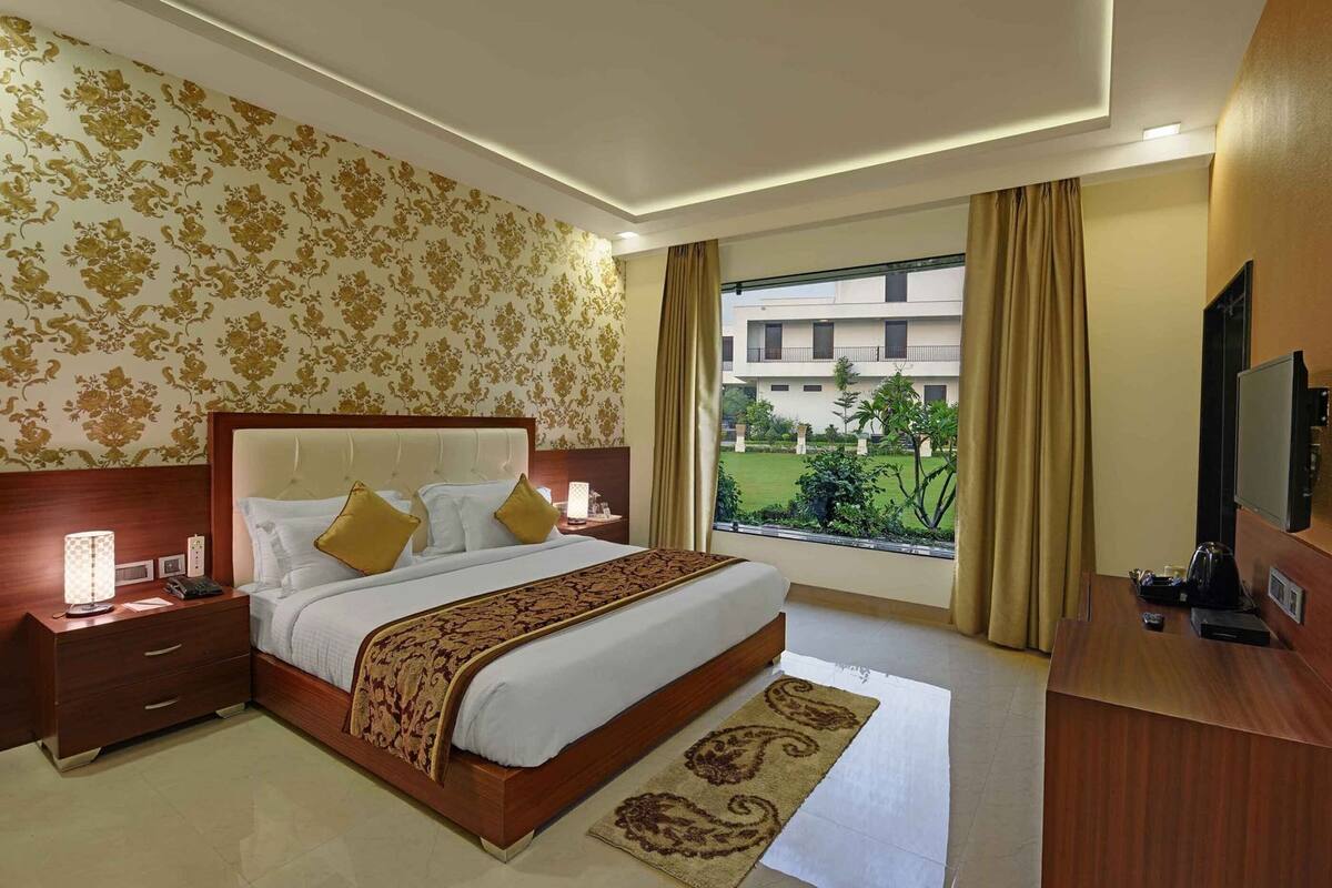 A hotel room with a large bed, a bedside table with a lamp, a TV, and a view of a building and greenery outside.