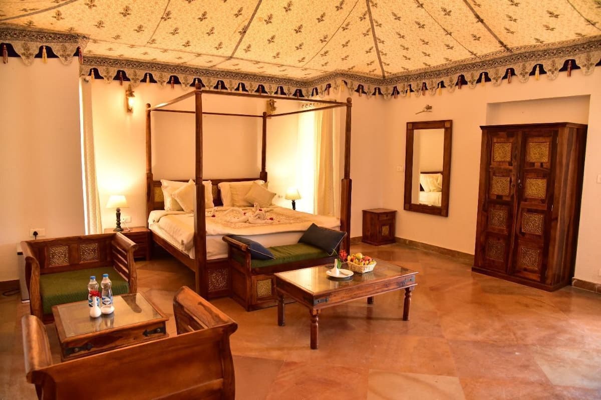 A four-poster bed with a green cushion, a wooden table with a fruit basket, and a wooden cabinet.