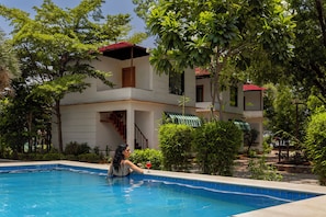A swimming pool with a person sitting by the edge, surrounded by lush greenery and a two-story building with a red roof.