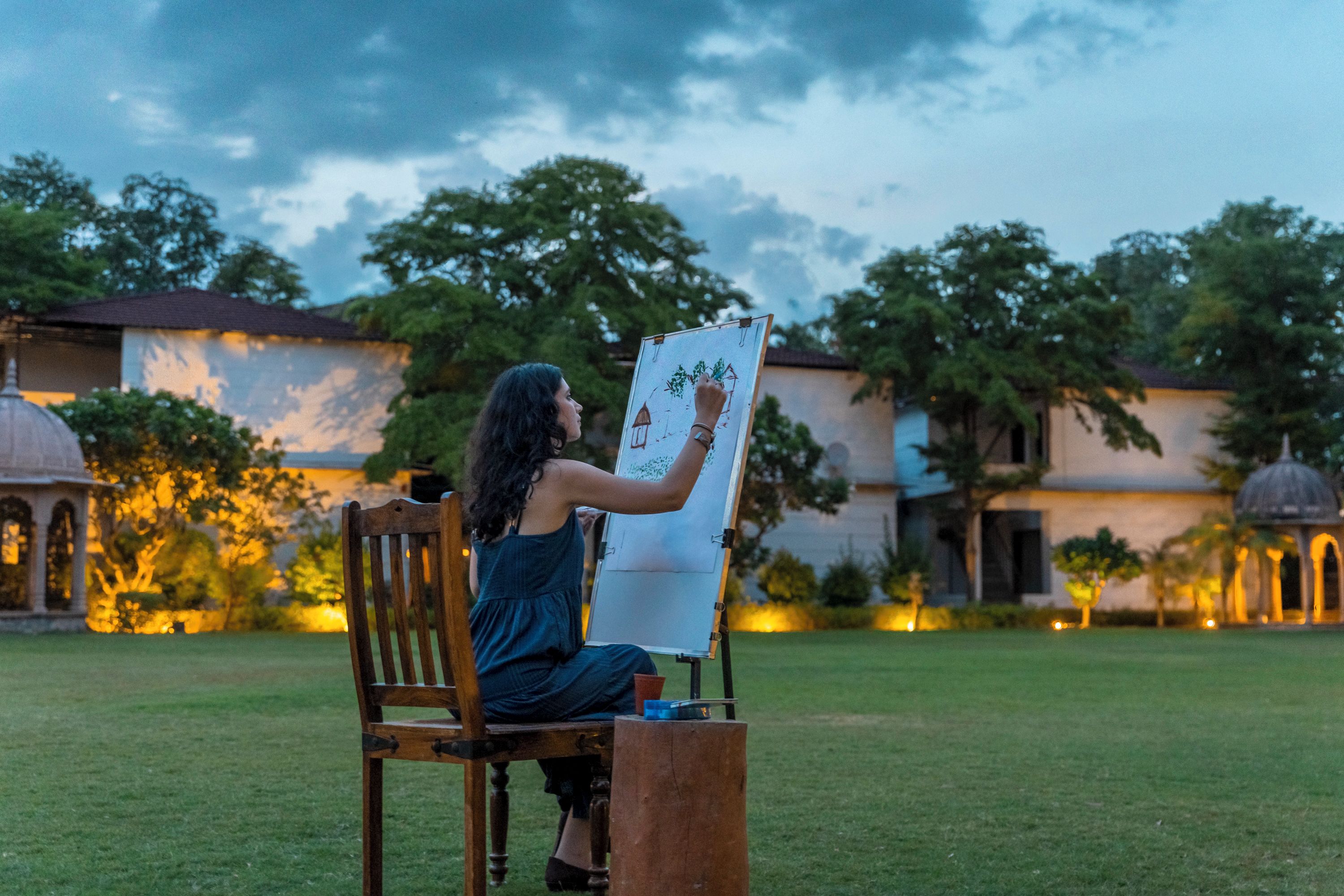 A person seated on a wooden chair, drawing on a whiteboard outdoors with a building and trees in the background.