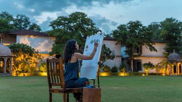 A person seated on a wooden chair, drawing on a whiteboard outdoors with a building and trees in the background.