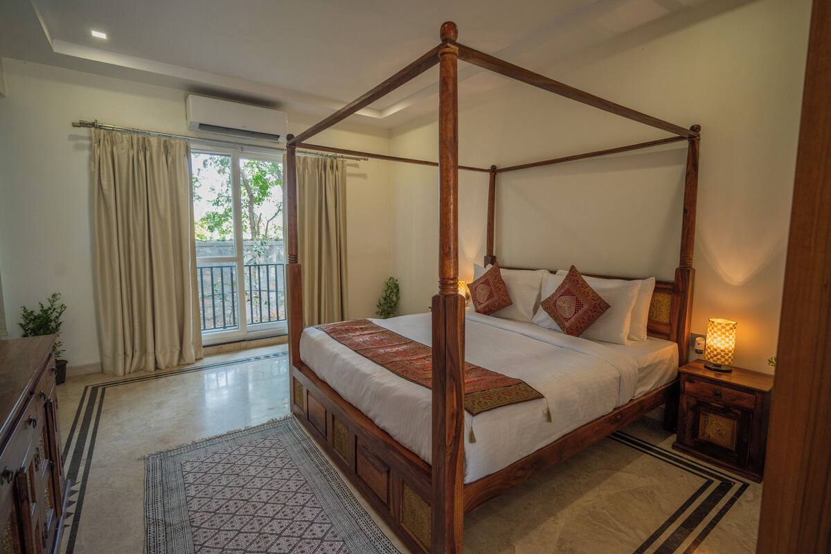 A four-poster bed with a wooden frame, a bedside table with a lamp, and a view of the outdoors through a sliding glass door.