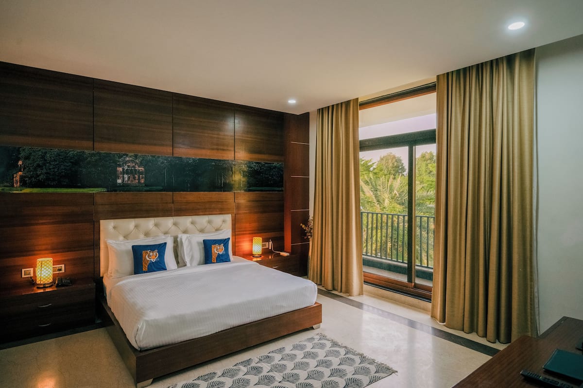 A modern bedroom with a large bed, wooden paneling, a nightstand, a bedside table, and a view of greenery through a large window.