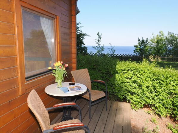Una cabaña de madera con un porche, una mesa con sillas, un florero con flores y vistas al mar.