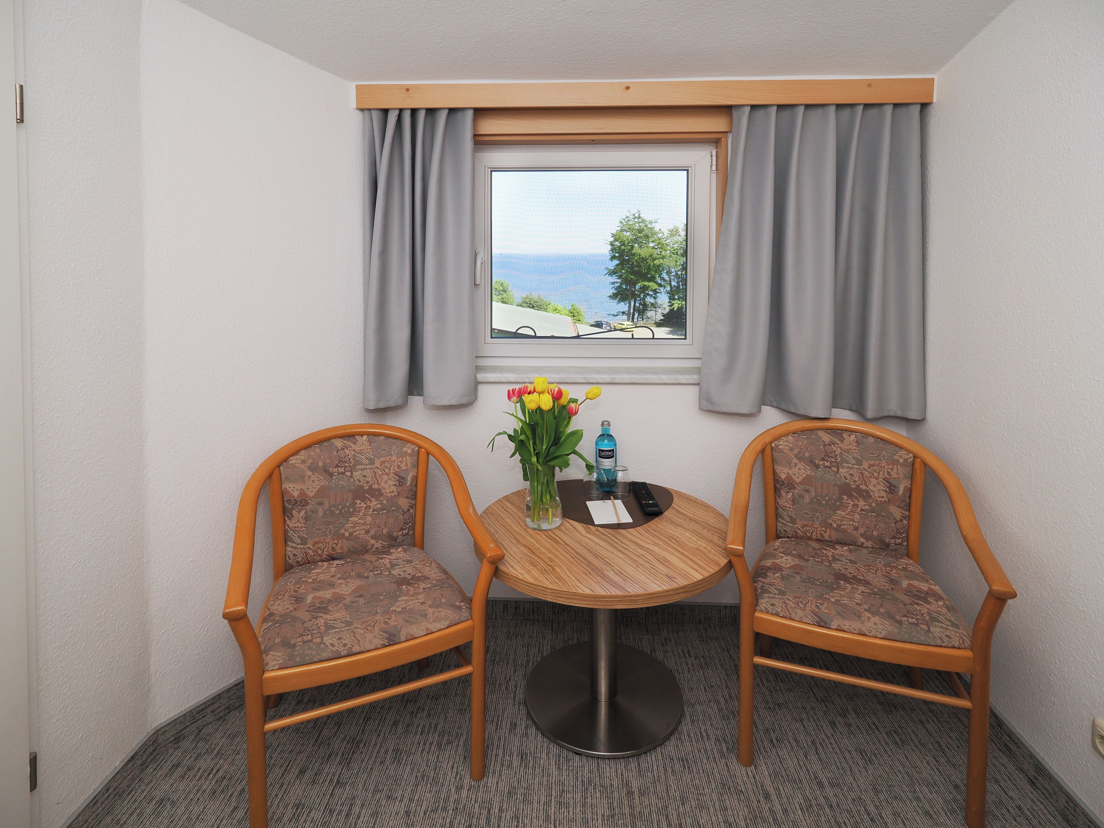 A small room with two wooden chairs, a round table, a vase with flowers, and a window with curtains.