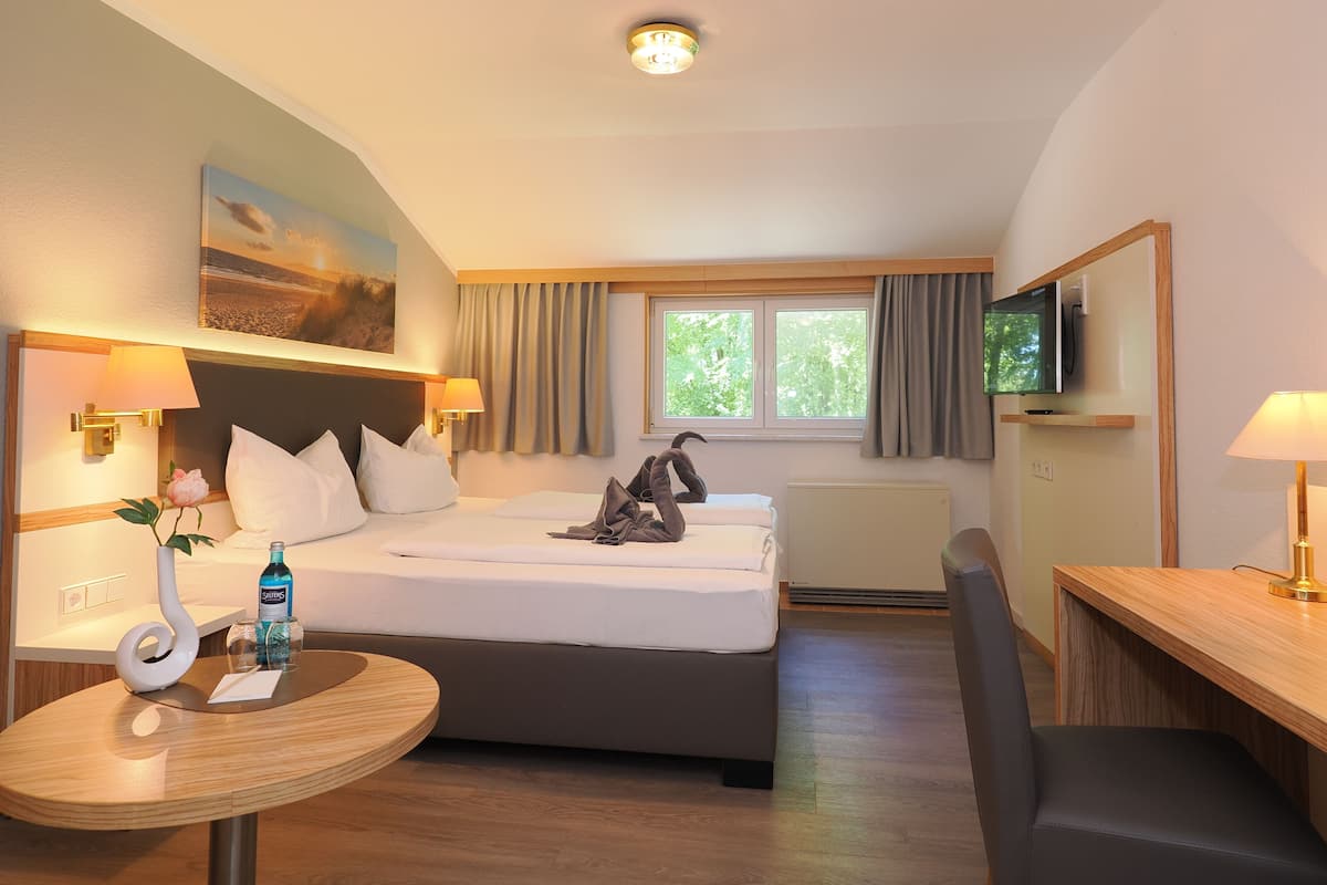 A hotel room with a bed, bedside table, chair, desk, lamp, TV, and a window with curtains.