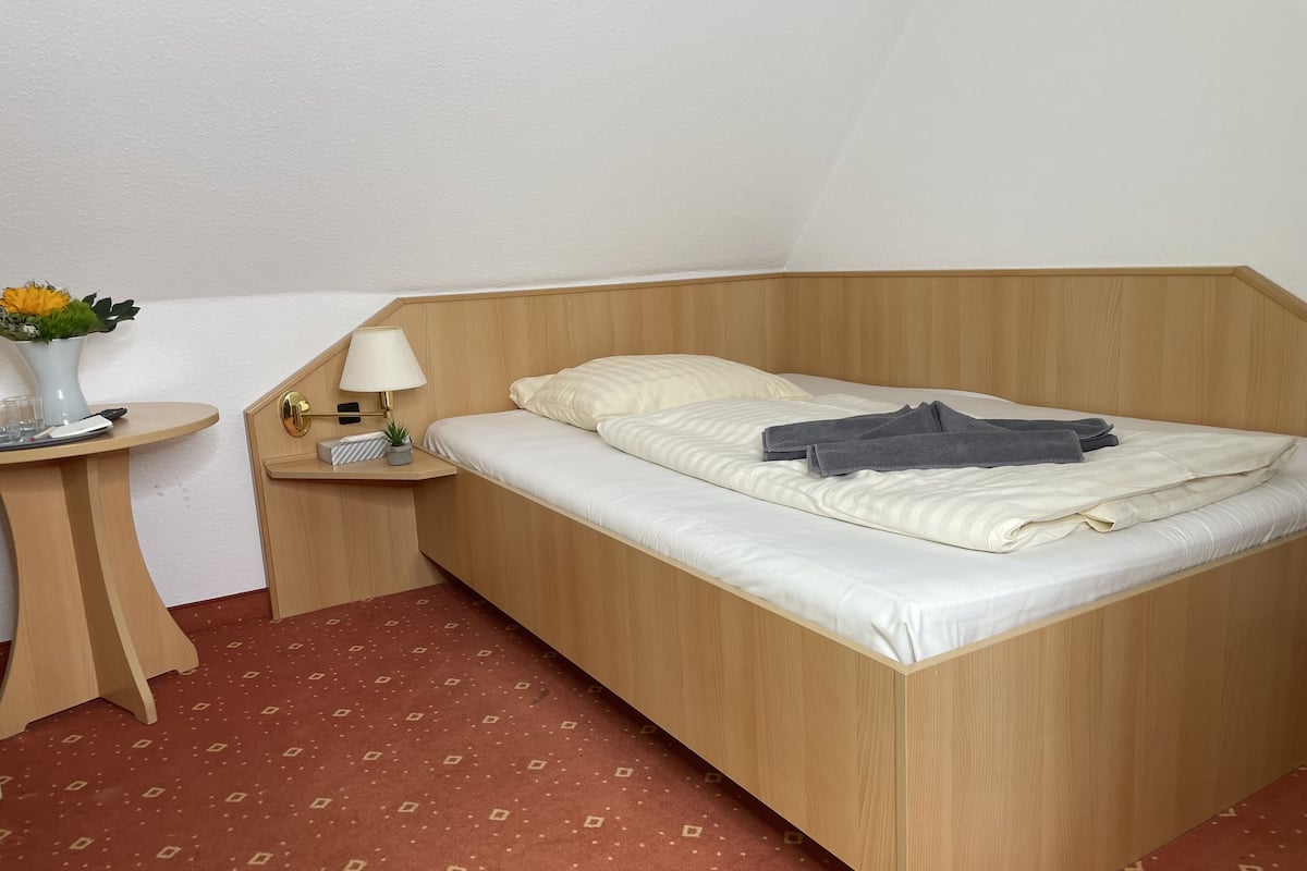 A single bed with a wooden headboard, a bedside table with a lamp, a small plant, and a vase with flowers.