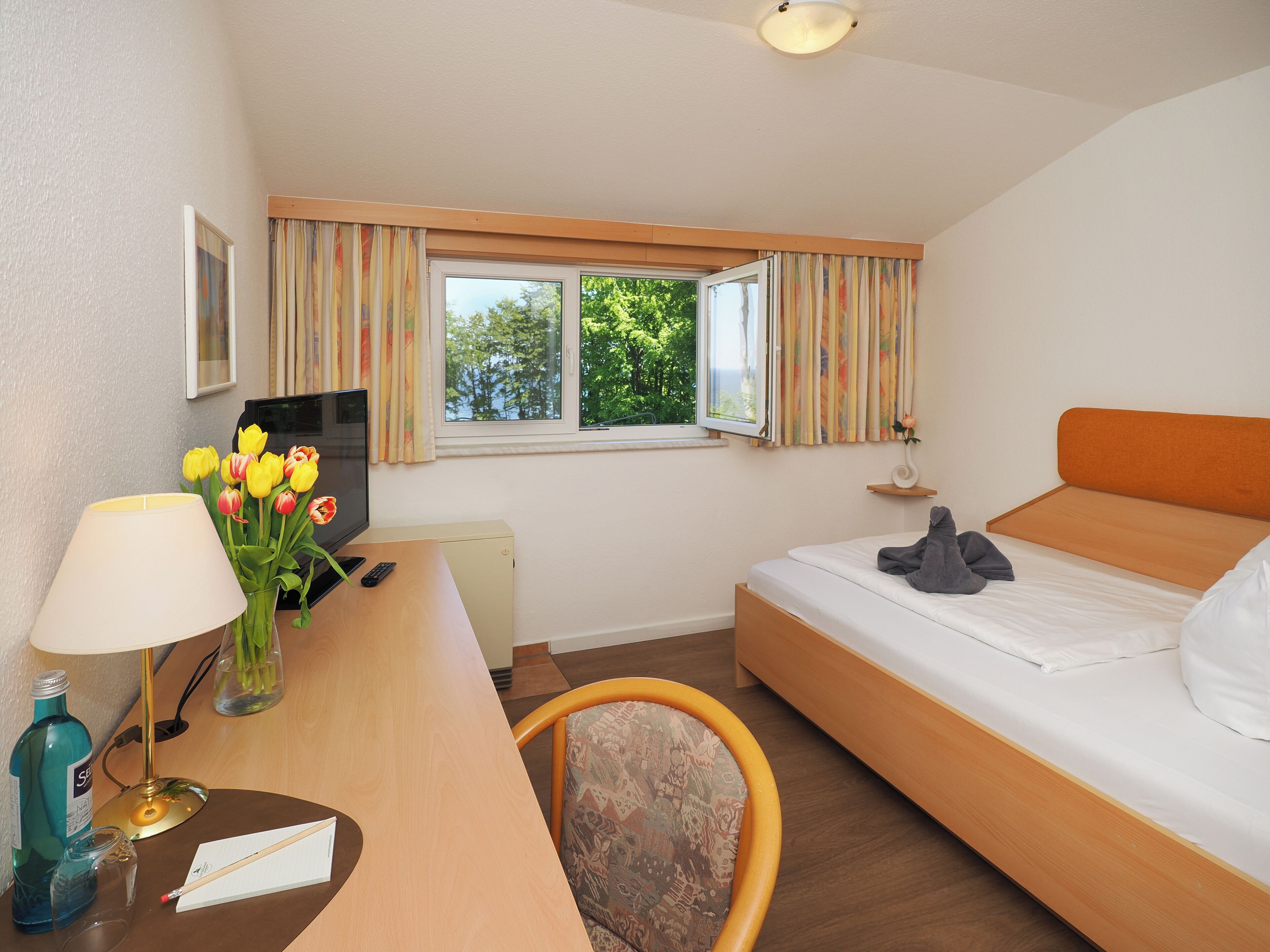 Une chambre d’hôtel avec un lit, un bureau, une chaise, une télévision, une lampe, un vase avec des fleurs et une fenêtre avec des rideaux.