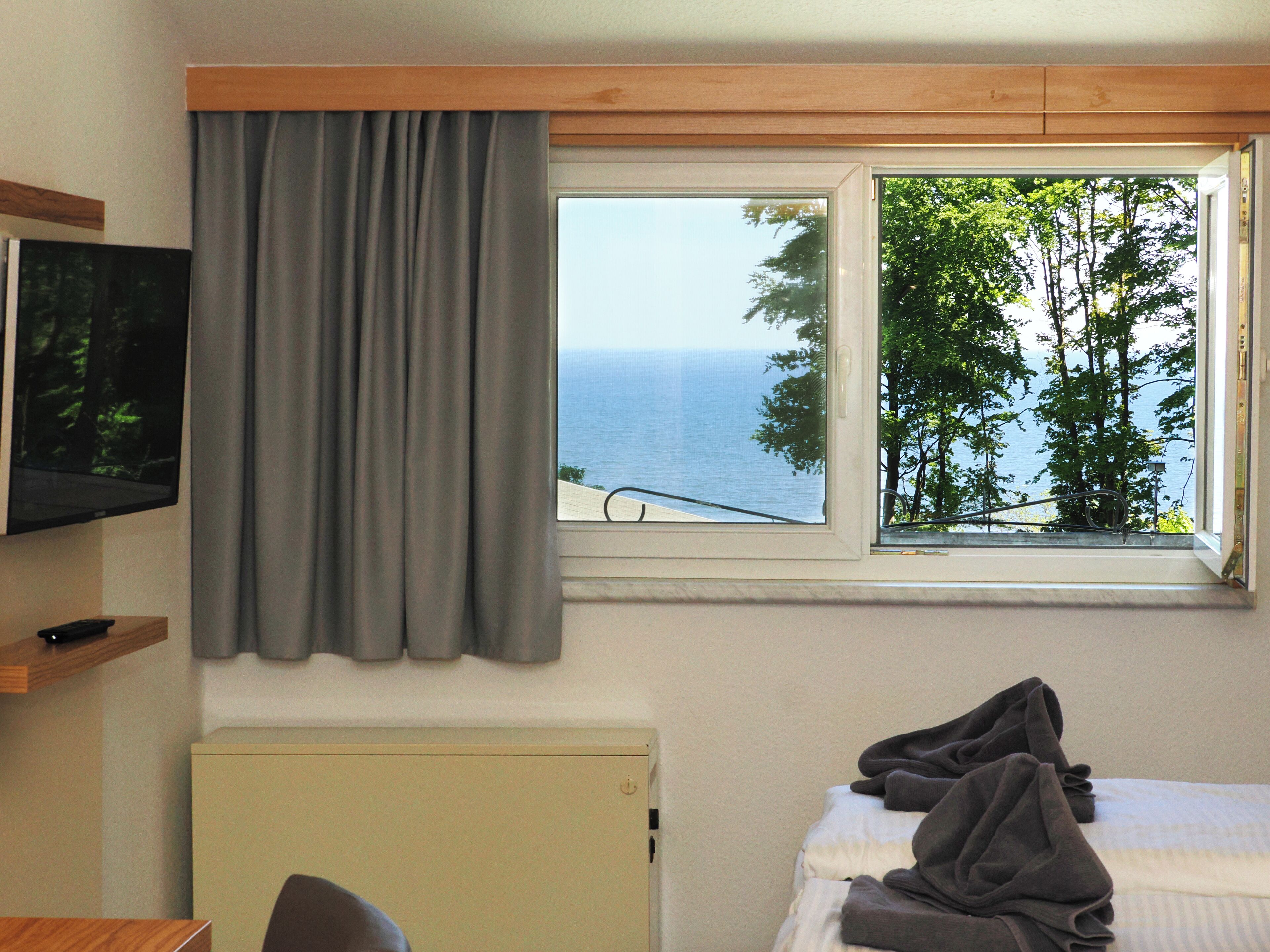 A room with a bed, a TV mounted on the wall, a window with a view of the sea and trees, and a radiator.