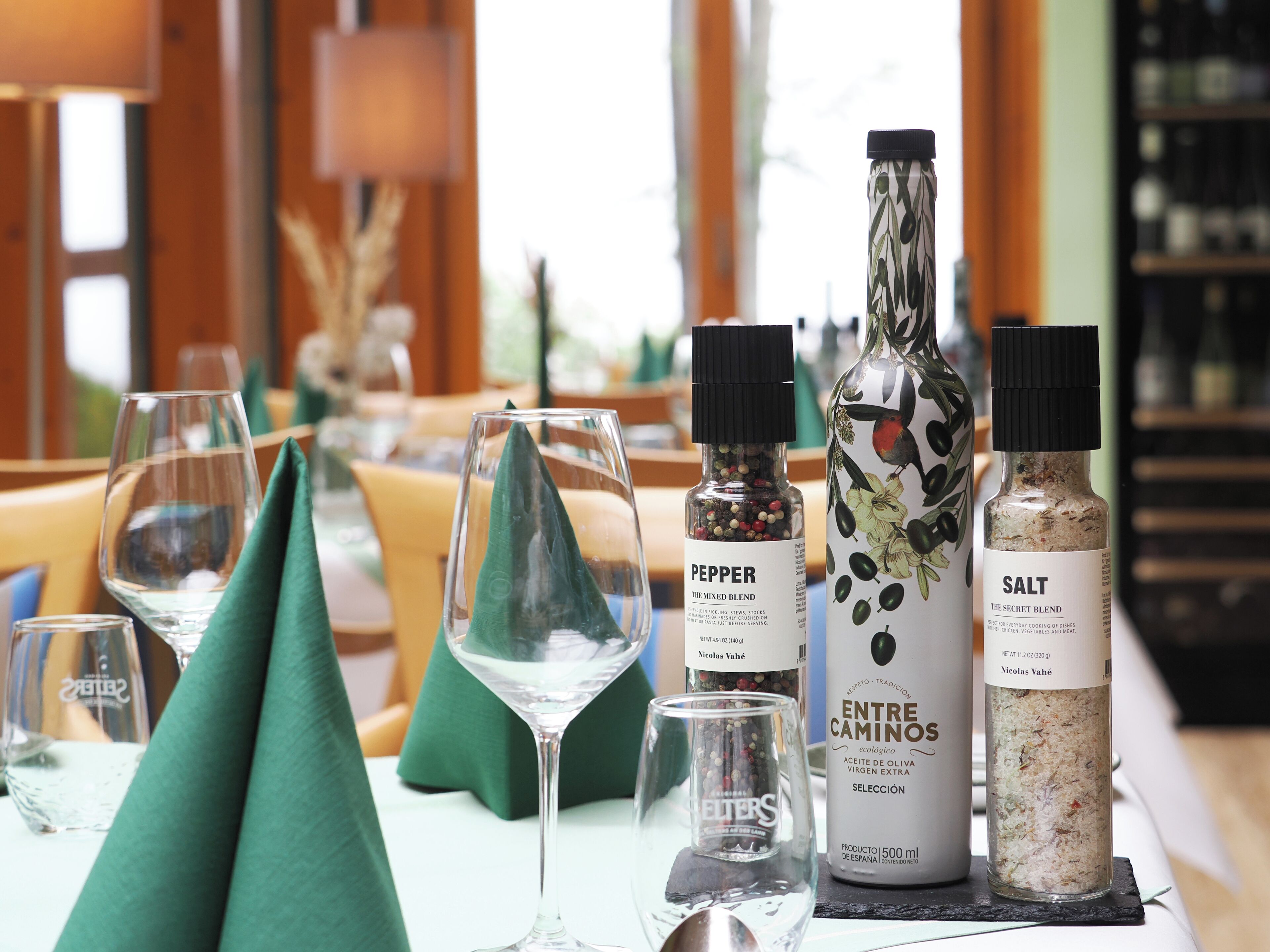 Una mesa servida con copas de vino, un mantel verde y tres botellas de condimentos etiquetadas como 'Pepper', 'Entre Caminos' y 'Salt'.