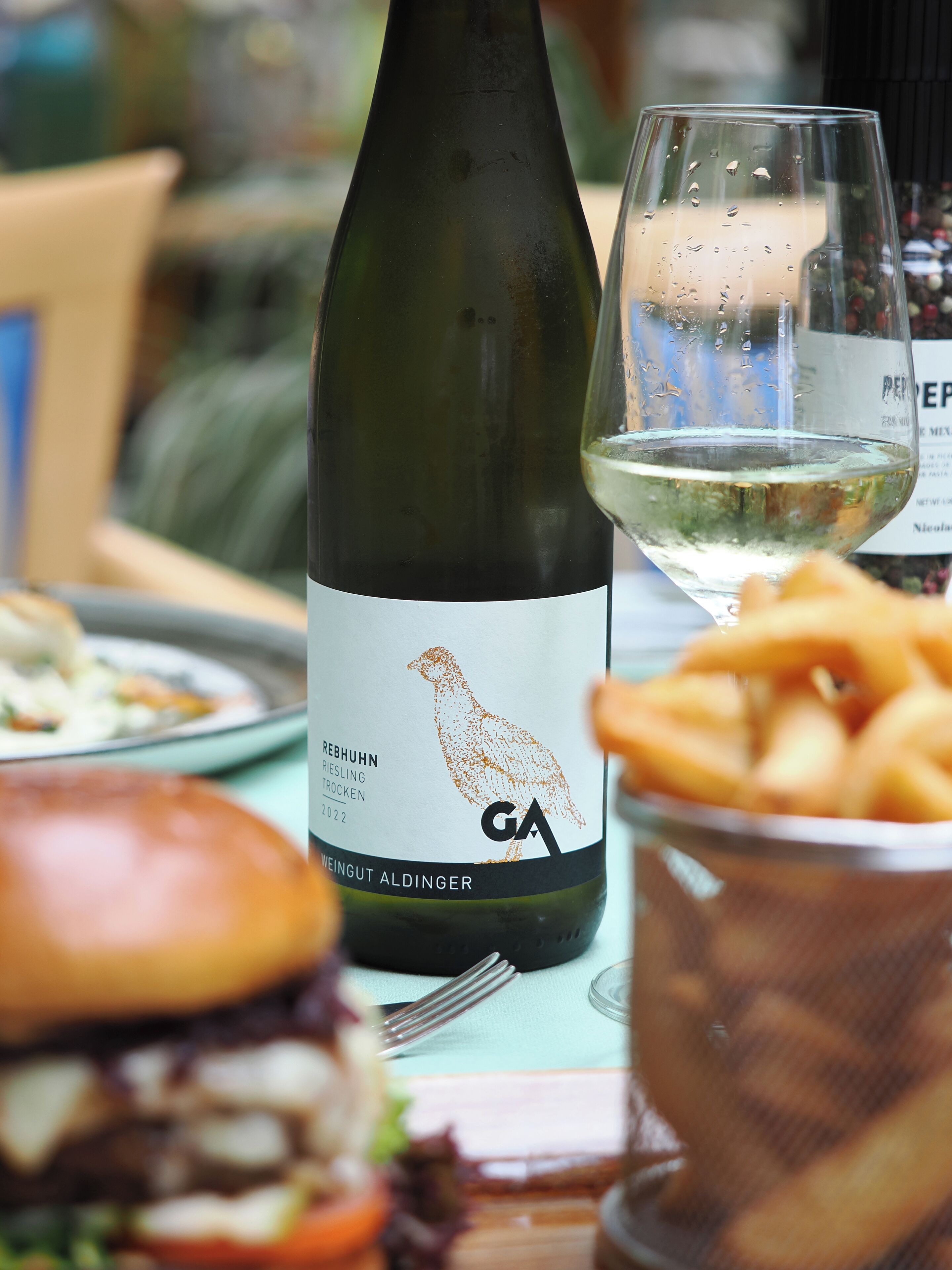 A bottle of wine with a label, a glass of white wine, a burger, and a bowl of fries on a table.