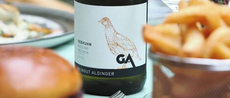 A bottle of wine with a label, a glass of white wine, a burger, and a bowl of fries on a table.