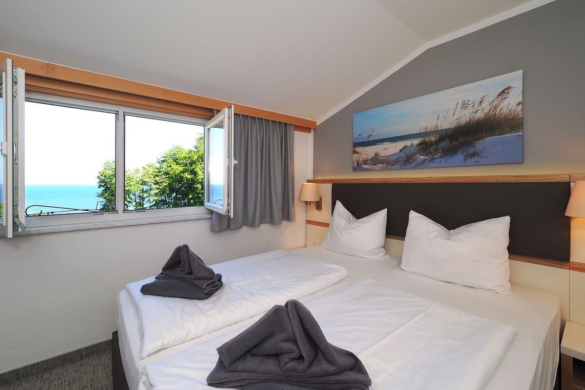 A hotel room with two beds, a large window with a view of the sea, and a painting on the wall.