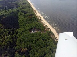 Vista aérea de una zona costera con bosque denso, una playa de arena y un pequeño edificio con paneles solares.