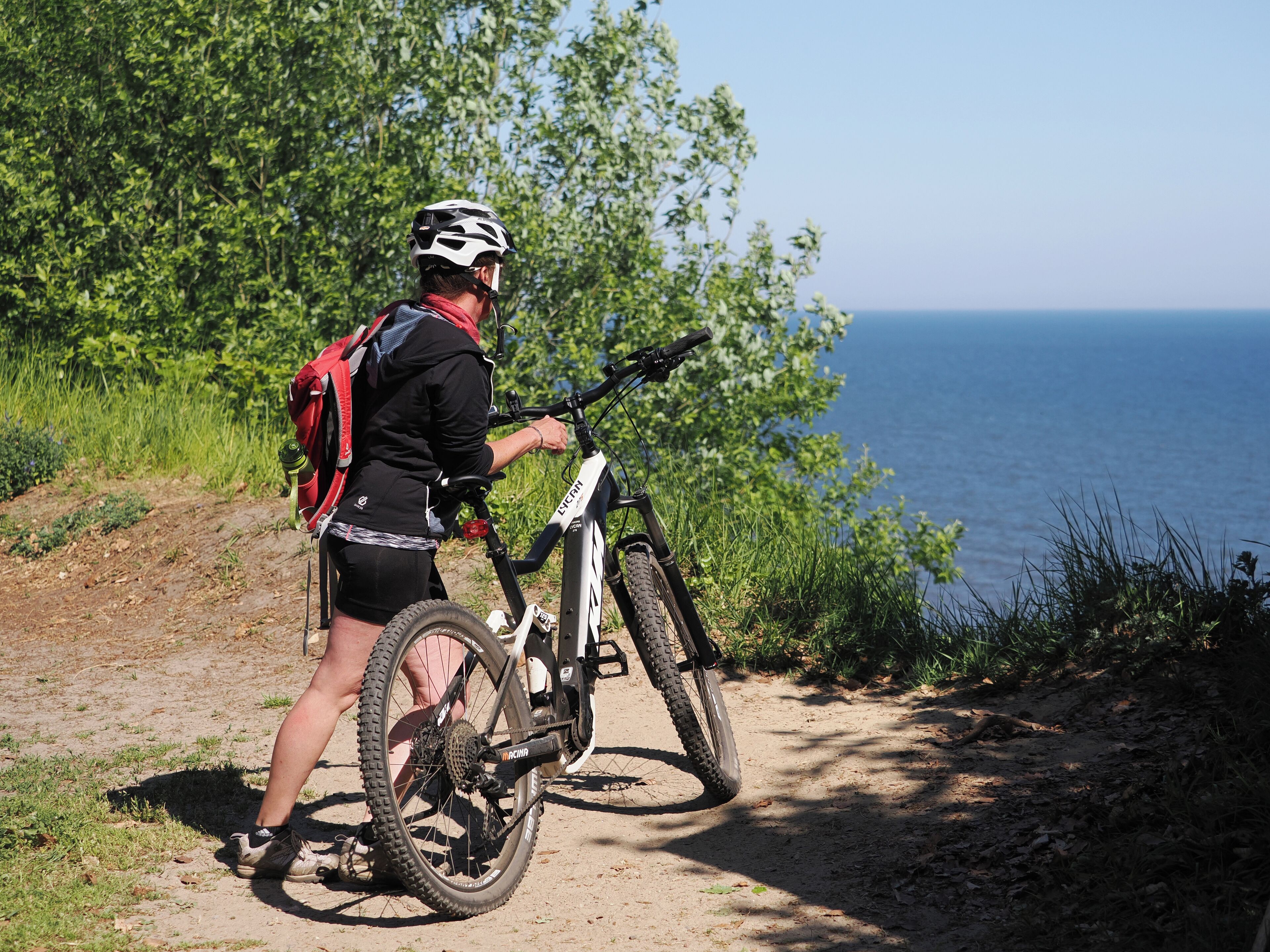 A person standing next to a mountain bike on a dirt path with greenery and a body of water in the background.